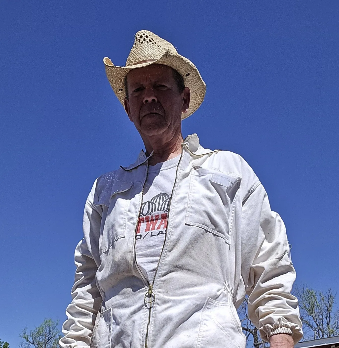 Big Dummy, the bee wrangler, standing in a straw hat and white bee jacket against a clear Texas sky