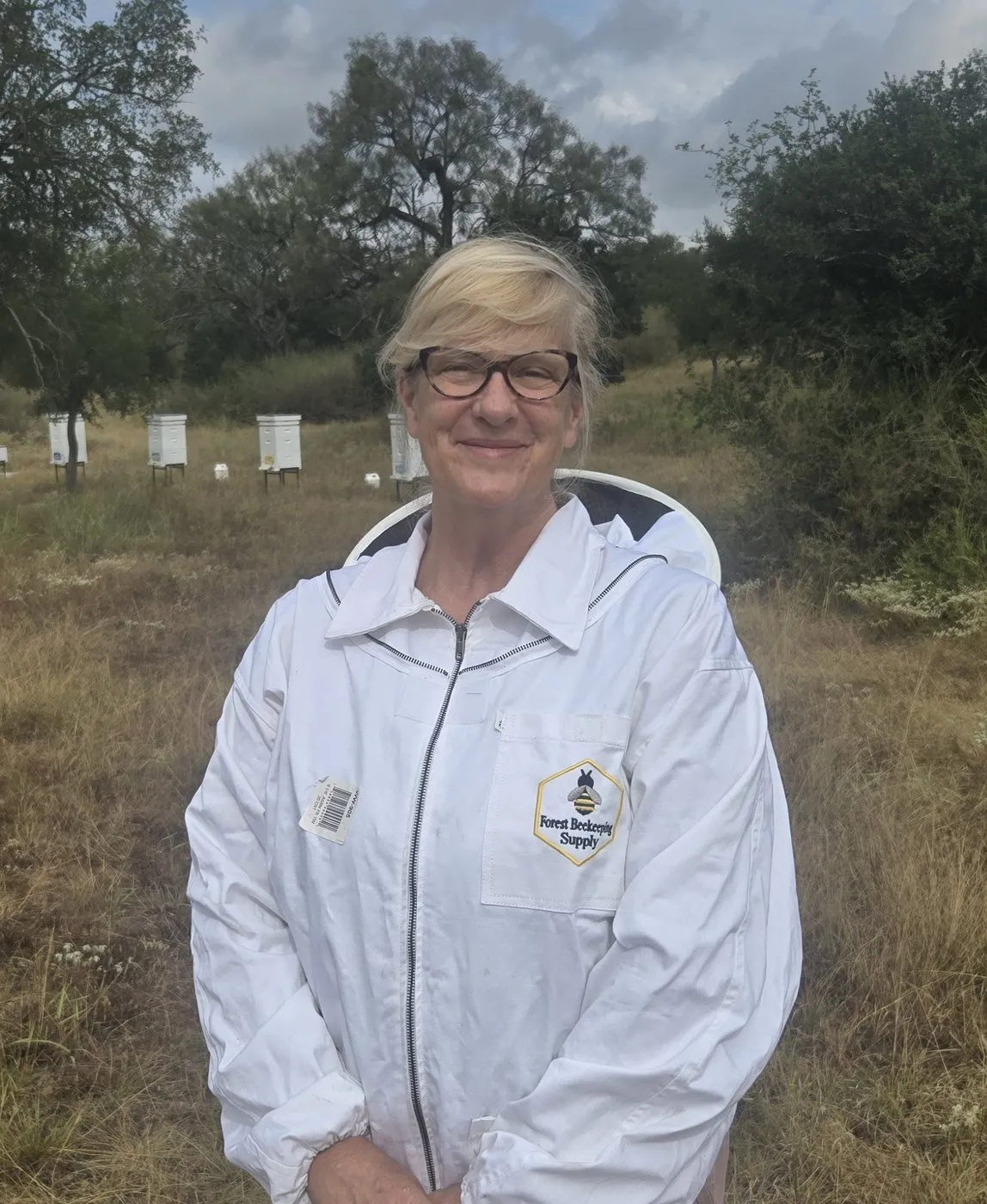 Miss Honey, The Boss, in a white bee jacket with Hill Country hives and live oak behind her